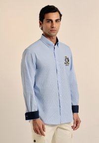 Camisa a rayas azul claro con mangas largas, cuello abotonado, puños azul marino y un logo bordado en el pecho.