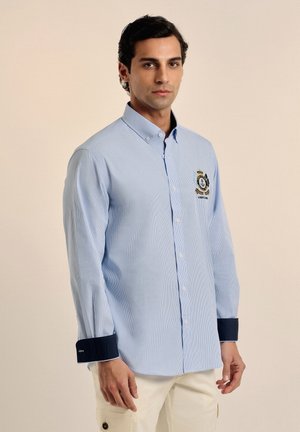 Camisa a rayas azul claro con mangas largas, cuello abotonado, puños azul marino y un logo bordado en el pecho.