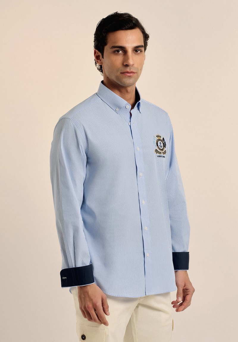 Camisa a rayas azul claro con mangas largas, cuello abotonado, puños azul marino y un logo bordado en el pecho.