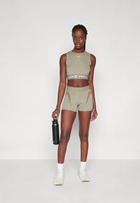 Set sportivo verde oliva composto da un top corto e pantaloni shorts con logo "Nike Pro". Il modello tiene in mano una bottiglia d'acqua nera; indossa delle sneakers bianche.