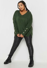 Green, oversized V-neck sweater with side slits and decorative buttons on the sleeves, paired with black faux leather pants and boots.