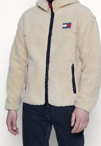 Uomo che indossa una giacca in sherpa beige con zip nera e patch del logo Tommy Jeans, abbinata a jeans blu scuro.