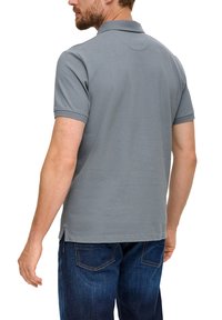 Polo gris avec un col structuré, des manches courtes et des fentes sur les côtés. Le tissu présente une finition texturée, associé à un jean foncé.