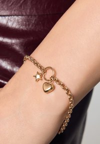 Złota bransoletka z łańcuchem, ozdobiona zawieszkami w kształcie gwiazdy i serca, o polerowanym wykończeniu i splatanych ogniwach, noszona na nadgarstku.