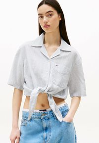 Camisa de botones de manga corta, color azul claro, con un lazo en la parte delantera. Hecha de una tela texturizada, presenta un bolsillo en el pecho y rayas sutiles.