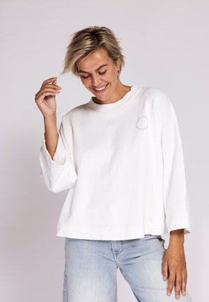 Woman with short blonde hair wearing a loose white long-sleeve top and light blue jeans smiling and looking down with one hand raised.