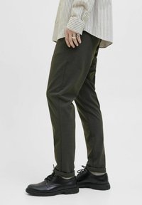 Pantaloni strâmți de culoare verde închis, cu un finisaj texturat, asortați cu pantofi negri cu șireturi. Buzunarele pantalonilor sunt ușor rulate, dezvăluind șosete.