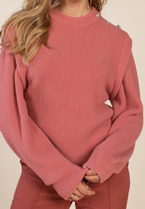 Femme portant un pull côtelé rose poussiéreux avec des boutons aux épaules, manches longues et mains jointes devant, associé à un pantalon couleur rouille.
