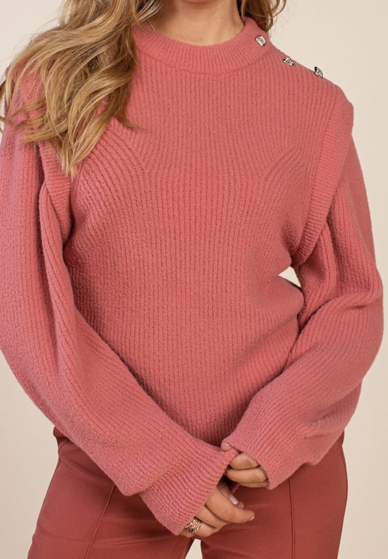 Femme portant un pull côtelé rose poussiéreux avec des boutons aux épaules, manches longues et mains jointes devant, associé à un pantalon couleur rouille.