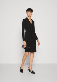 Svart klänning med långa ärmar, v-ringning och rynkdetaljer vid midjan. Bärs med svarta slingback-klackar och en strukturerad svart axelväska.
