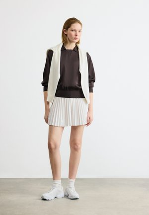 Mladá žena stojí oblečená v čiernom tričku s dlhým rukávom, bielej skladanej sukni, bielych teniskách a s bielym svetrom prehodeným cez ramená.