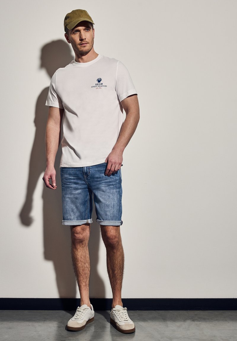 Vit bomulls t-shirt med blå tryckt grafik, i kombination med blå denimshorts och vita sneakers, vilket kompletterar en avslappnad sommarslook.