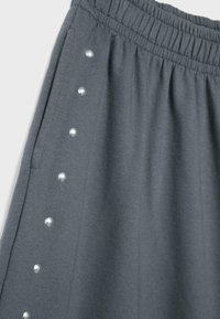 Pantalon à taille élastique grise avec une rangée latérale de boutons-pression argentés et une poche latérale, montrant la texture du tissu et les détails des coutures.