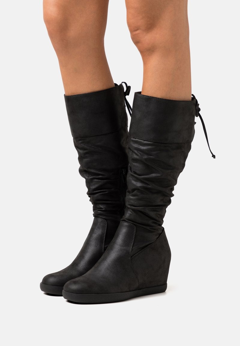 Bottes compensées noires mi-mollet avec revers plié et cordon à nouer à l'arrière, portées sur des jambes nues sur un fond blanc.