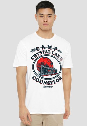 Young man wearing white t-shirt with "Camp Crystal Lake Counselor" text and cabin graphic in front, referencing Friday the 13th film.