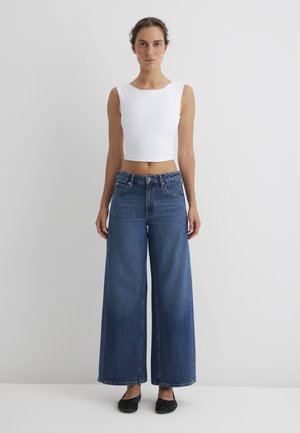 Weite blaue Jeans aus Denim mit hoher Taille, kombiniert mit einem taillierten, weißen ärmellosen Oberteil. Schwarze geflochtene Slipper runden das Outfit ab.