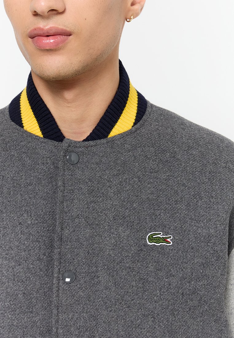 Gros plan sur une personne portant une veste grise avec un col côtelé jaune et marine et un petit logo de crocodile vert sur la poitrine.