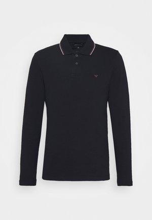 Pikkade varrukatega must polo särk, millel on kaelus punaste ja valgete triipudega, kolmeknöpplihe, ning väike broderi logo rinnal.