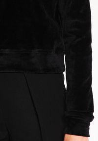 Persona che indossa un top in pelliccia nera a maniche lunghe e pantaloni neri, mostrando il busto e il braccio su uno sfondo bianco.