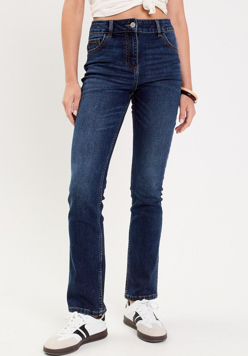 Jeans high-rise blu scuro in denim con cinque tasche, cuciture a vista e una vestibilità slim, abbinati a sneakers bianche e grigie.