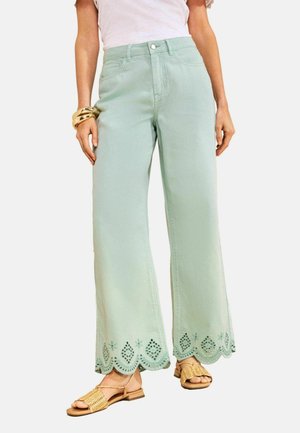 Woman wearing light green wide-leg pants with scalloped, embroidered hems, a white top, gold bangles, and woven tan sandals.
