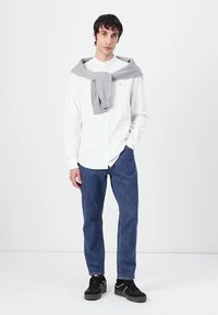 Weißes Hemd mit Knöpfen, graue Strickjacke über den Schultern, blaue Jeans und schwarze Sneakers an einem Modell vor einem neutralen Hintergrund.