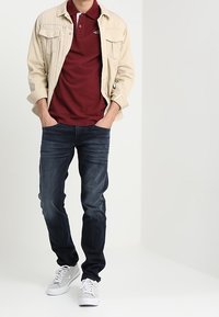 Béžová denimová bunda přes vínově červené polo tričko, doplněná tmavě modrými džíny a bílými teniskami. Bunda má dvě náprsní kapsy.