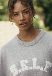 Maglione grigio a maglia con colletto tondo, con lettering in rilievo bianco che forma la scritta "S.E.L.F." sul davanti, indossato su uno sfondo naturale sfocato.