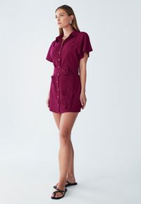 Söötud magenta värvi kleit, millel on lühikesed varrukad, nupclosure ja kaks esikäe taskut. Sile kangas ja kohandatud disain.