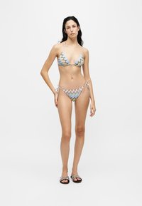 Femme debout portant un bikini à motif zigzag pastel avec des liens sur les côtés et des sandales slides assorties sur un fond clair uni.