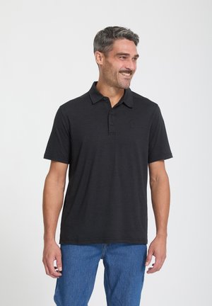 Lächelnder Mann mit kurzen grauen Haaren, der ein schwarzes Poloshirt und blaue Jeans trägt, steht vor einem einfarbigen weißen Hintergrund.