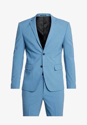 Svetlo modrý oblek vyrobený z textúrovanej tkaniny, s priliehavým strihom, dvojitým zapínaním, výstrihom s klopami a bočnými vreckami.