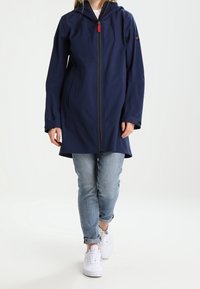 Person wearing a navy hooded zip-up jacket, light blue rolled-up jeans, and white sneakers, standing against a plain light background.