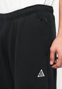 Sorte sweatpants med snøre i livet, sidelommer og et lille hvidt trekantlogo. Stoffet synes blødt med en struktureret overflade.