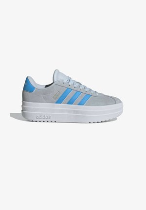 Sneaker in suede grigio chiaro con accenti blu, dotato di un design a tre strisce, una suola bianca spessa e punta rotonda. Dettaglio di branding minimale sul tallone.