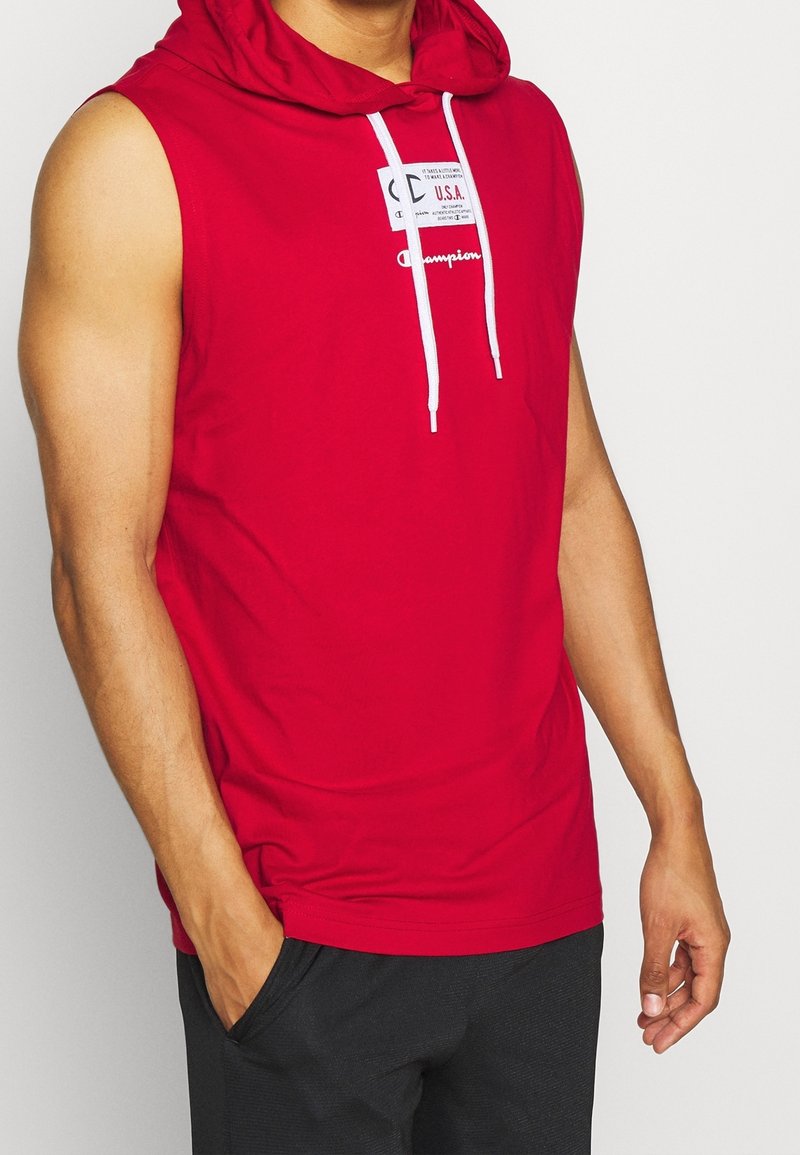 Punainen hihaton huppari, jossa on etuosassa logoplässi, valkoiset kiristysnauhat ja rento istuvuus. Kangas vaikuttaa sileältä ja kevyeltä.