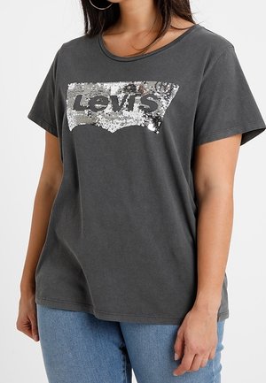 Femme portant un t-shirt Levi's gris foncé avec un logo en sequins argentés et un jean bleu clair, debout devant un fond blanc uni.