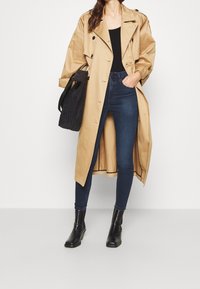 Long beige trench coat with black buttons over a black top and dark blue jeans. Black ankle boots, holding a textured black bag.