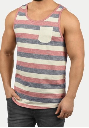 Une personne musclée porte un débardeur sans manches à rayures horizontales rouges, grises et blanches avec une poche poitrine blanche, associé à un jean foncé.