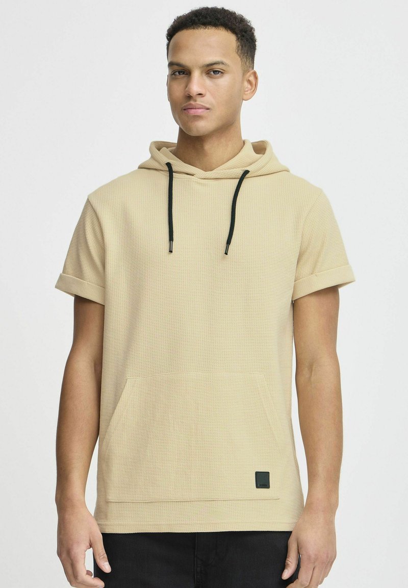Beige kortærmet hætte sweatshirt med et tekstureret mønster, forlommer, sorte snore og et lille sort logo-patch ved kanten.