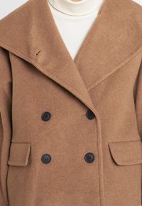 Brun ullkappa med en stor slagkrage, dubbelknäppta svarta knappar och två framfickor. Texturen verkar mjuk och varm.