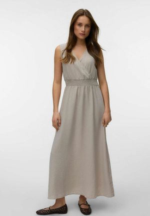 Kobieta stoi, ubrana w bezrękawową beżową sukienkę maxi z marszczonym stanem i brązowe plecione baleriny na jednolitym białym tle.