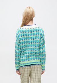 Maglione lavorato a maglia in tonalità di blu e verde con motivi floreali, orlo a coste e colletto a contrasto. Indossato con pantaloni a quadri.