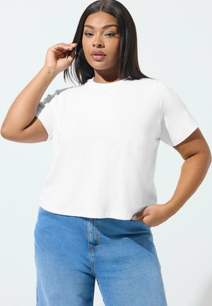 Kvinde med glat, mørkt hår iført en enkel hvid kortærmet skjorte og blå jeans, poserer med den ene hånd nær ansigtet og den anden på hoften.