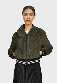 Olive zelená umelá kožuchová bomber bunda s predným zipsom, prehnaným golierom a pruhovanými rebrovanými okrajmi na spodku a na rukávoch. Model ju nosí cez plisovanú sukňu.