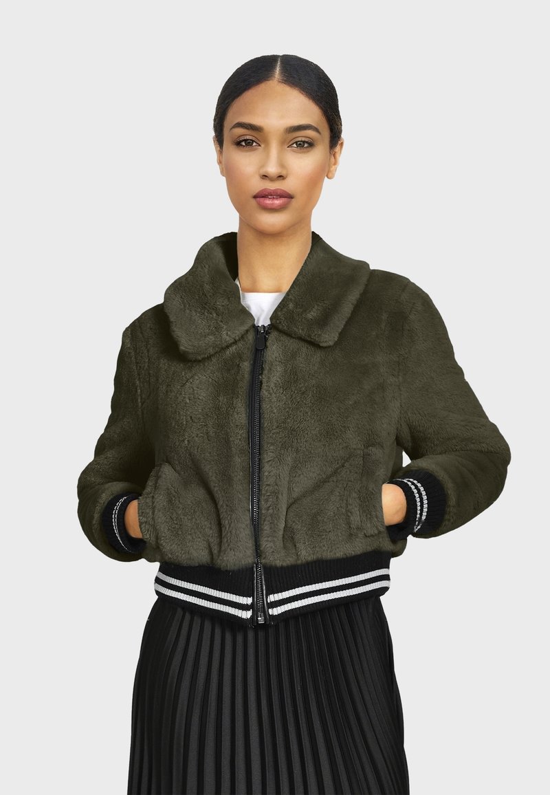 Olive zelená umelá kožuchová bomber bunda s predným zipsom, prehnaným golierom a pruhovanými rebrovanými okrajmi na spodku a na rukávoch. Model ju nosí cez plisovanú sukňu.