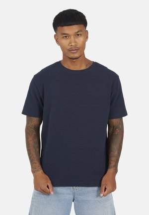 Uomo con capelli neri corti e braccia tatuate, indossa una t-shirt a maniche corte blu navy tinta unita e jeans azzurri chiari, su sfondo bianco.