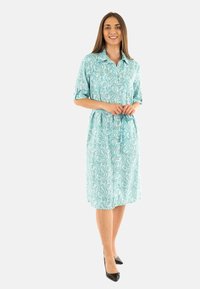 Un abito camicia azzurro chiaro con motivo a vortice, maniche corte, chiusura con bottoni e cintura in vita, abbinato a tacchi alti neri.