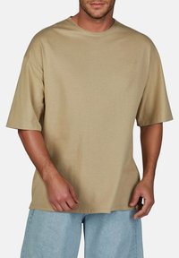 Beige katoenen t-shirt met een ronde halslijn en korte mouwen, met een losse pasvorm en een gladde textuur, gepresenteerd tegen een neutrale achtergrond.