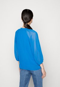 Blusa azul com mangas translúcidas e com bolinhas, decote arredondado e punhos elásticos. Usada com jeans azul claro, vista pelas costas.